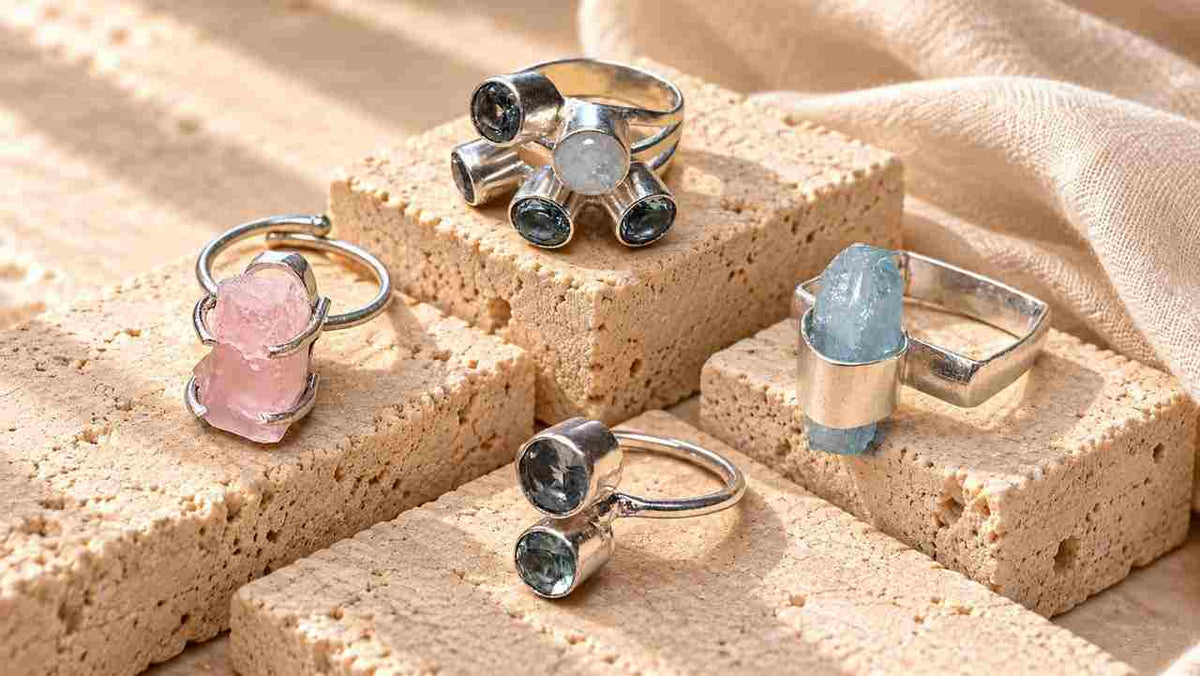 Anelli Quiet Stones con pietre naturali di colori e forme diverse, disposti su blocchi di travertino beige in una composizione editoriale luminosa.